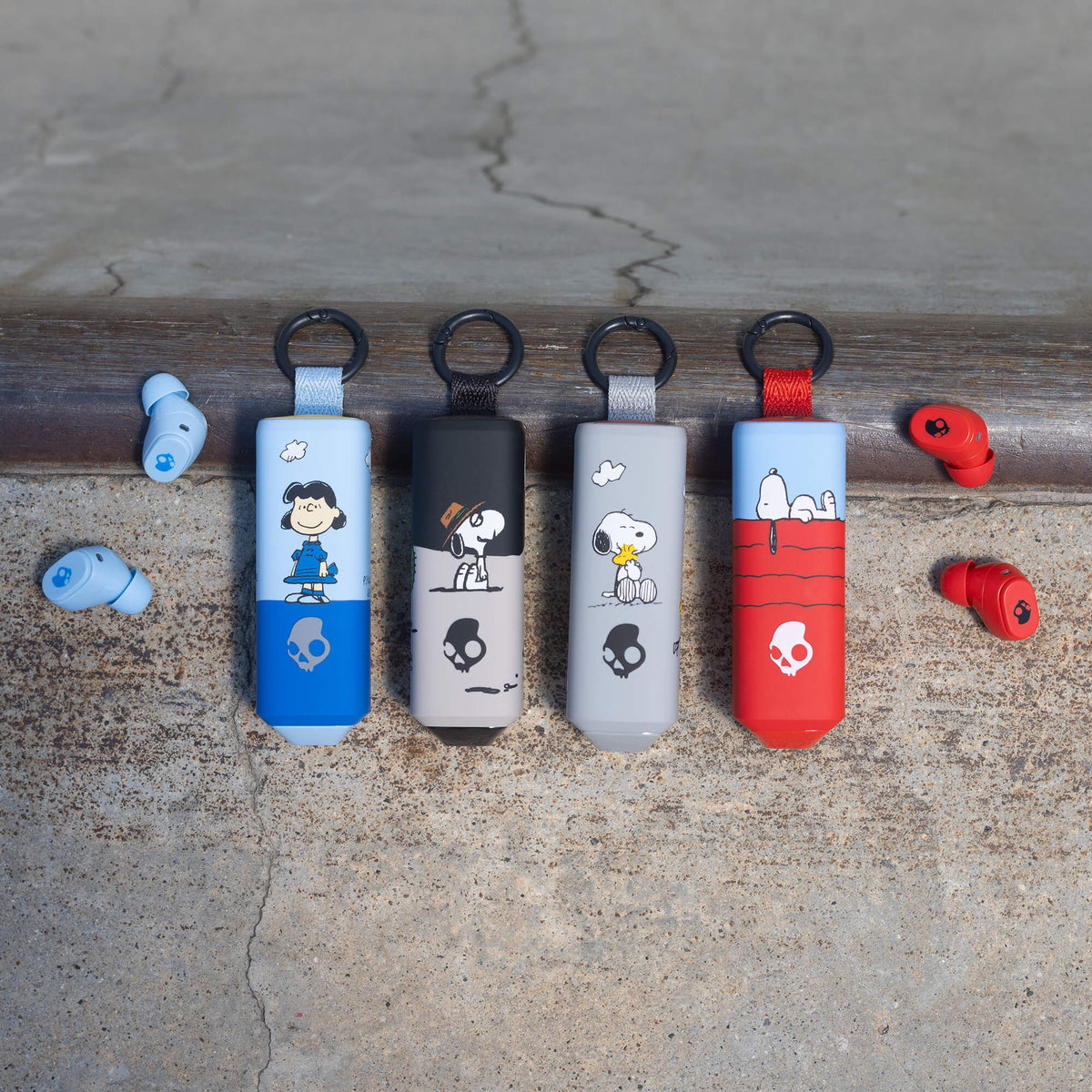Four Peanuts-themed dime 360 earbuds in blue, black, gray, and red are lined up on ramp coping, with matching red and blue earbuds resting on either side on a concrete slab. | Skullcandy X Peanuts Snoopy