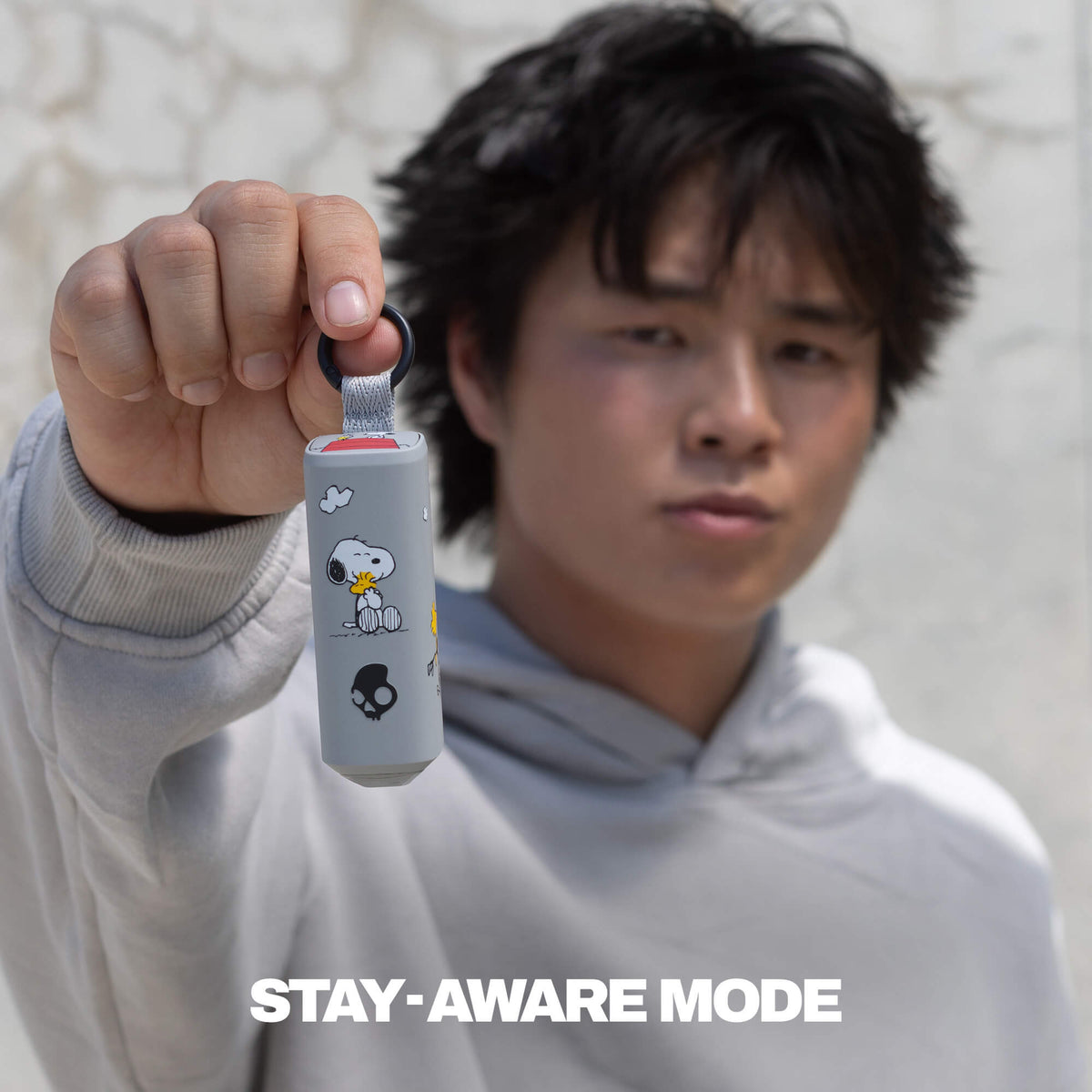 A person in a light gray hoodie holds up the dime 360 earbuds with woodstock-style cartoon graphics and a black loop at the top. The text 'STAY-AWARE MODE' appears at the bottom. | Skullcandy X Peanuts Woodstock