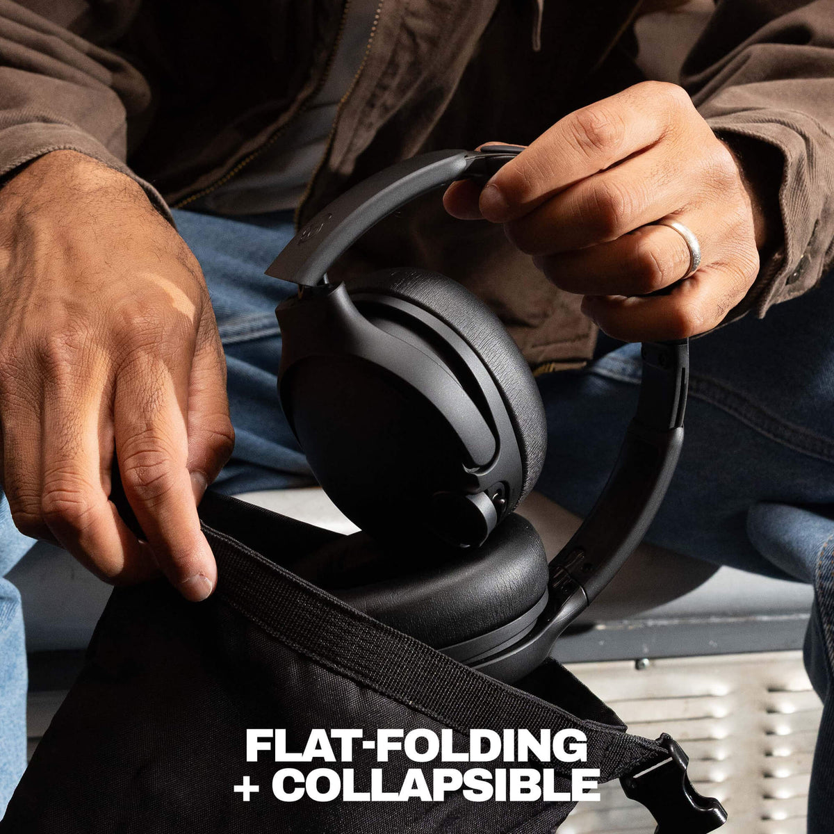A close-up of a person holding a black over-ear headset over an open black bag, with the text 'FLAT-FOLDING + COLLAPSIBLE' at the bottom. The person is wearing a brown jacket and blue jeans. | True Black